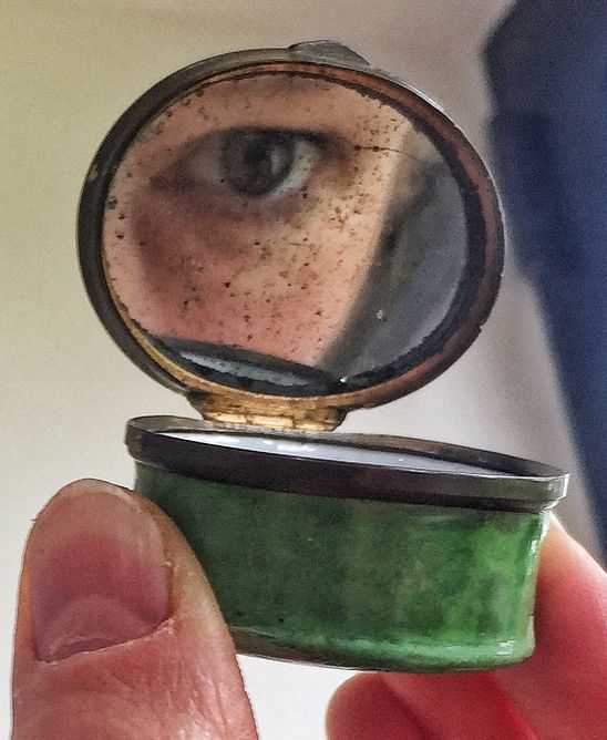 Fingers holding an enamel box with the lid open and an eye reflected in the mirror inside the lid.