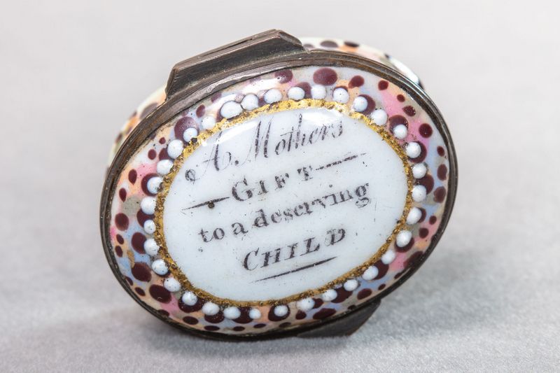 White, oval enamel box with a pink, brown and gold decorative border, including the typical white dots, and the motto in the centre.