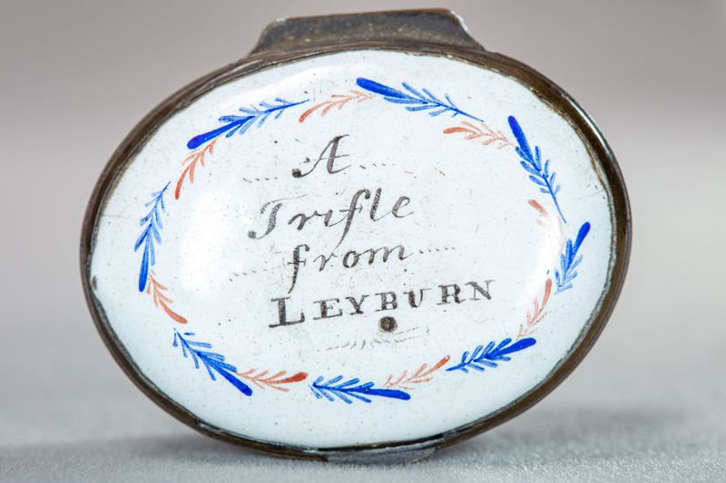 A very simple white enamel box with blue and red feathered patterned and the words in the middle