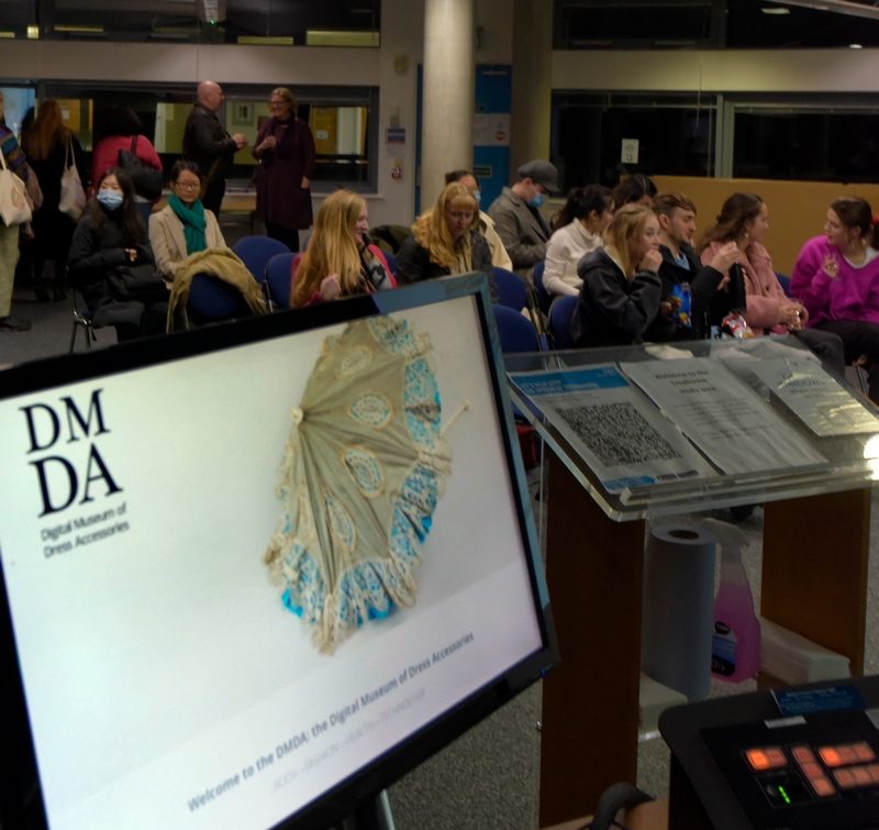 Computer screen in the foregoro0und showing the homepage of the DMDA website, with a room full of seated people beyond