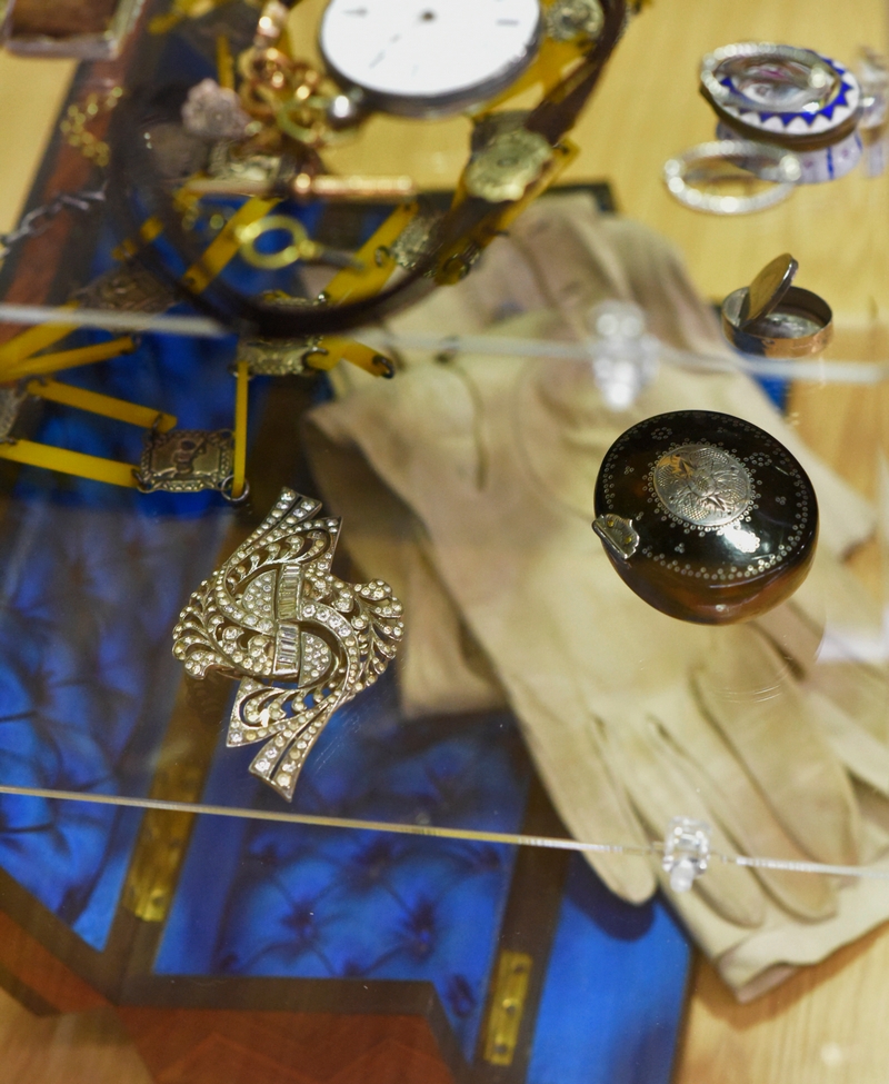 Closeup of an art-deco buckle and a patch box on a glass shelf; below them, blurred, is a pair of old kid gloves in an open, blue-silk-lined wooden box