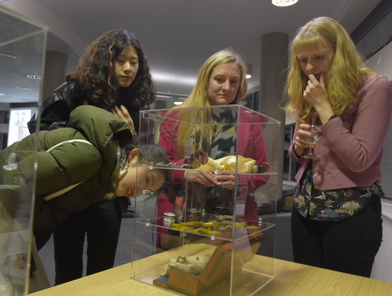 Four women standing around a small display cabinet on a table - one is bending down for a closer look